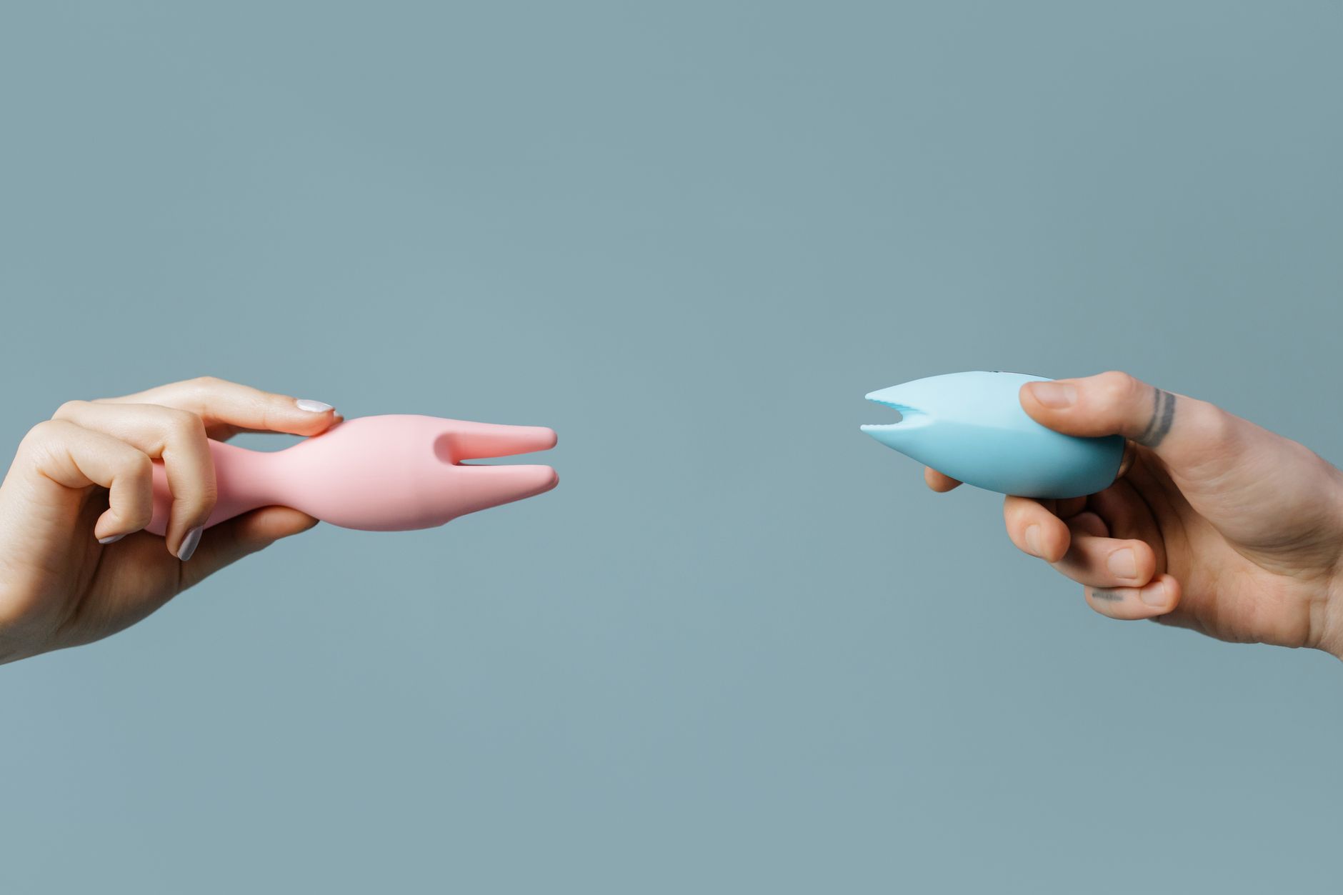Two hands holding pink and blue clitoral vibrators against a pastel background