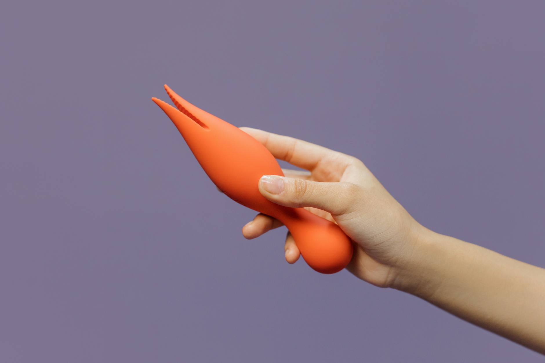 Close-up of a hand holding an orange vibrator against a minimalistic purple backdrop, showcasing modern sensuality.