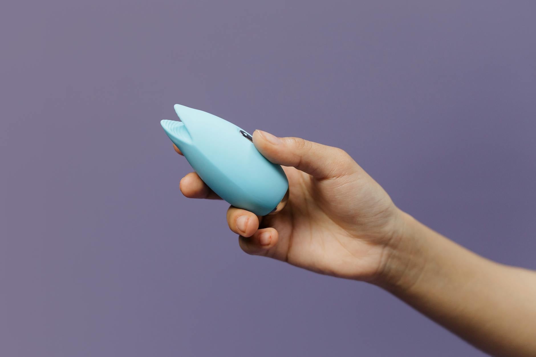 Two hands holding pink and blue silicone vibrators against a pastel background